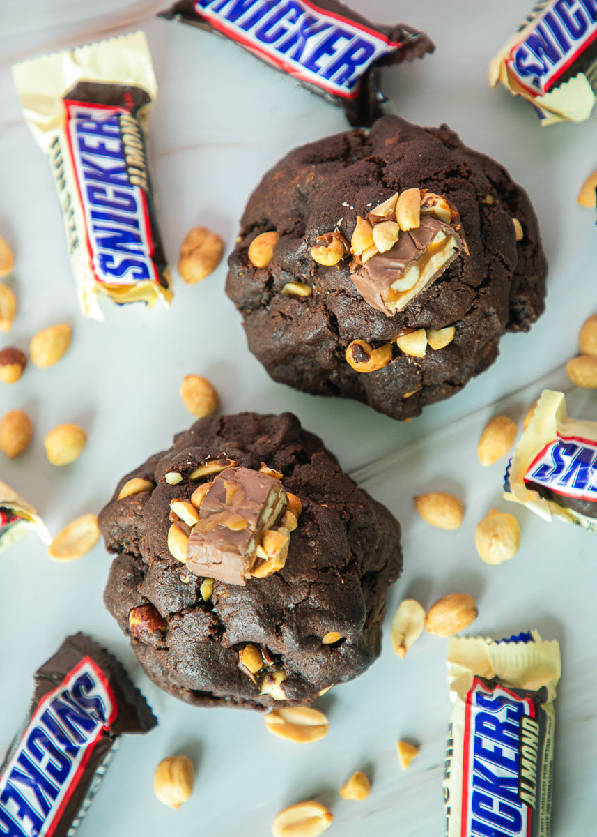 Chocolate cookies with Snickers pieces on a light blue background with scattered peanuts and Snicker bars.
