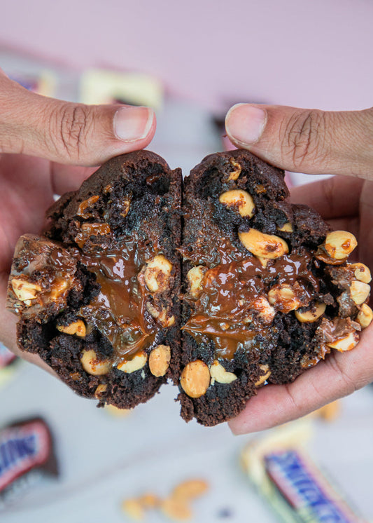 Chocolate cookie with peanut pieces held by a hand, with Snickers candy bars in the background.