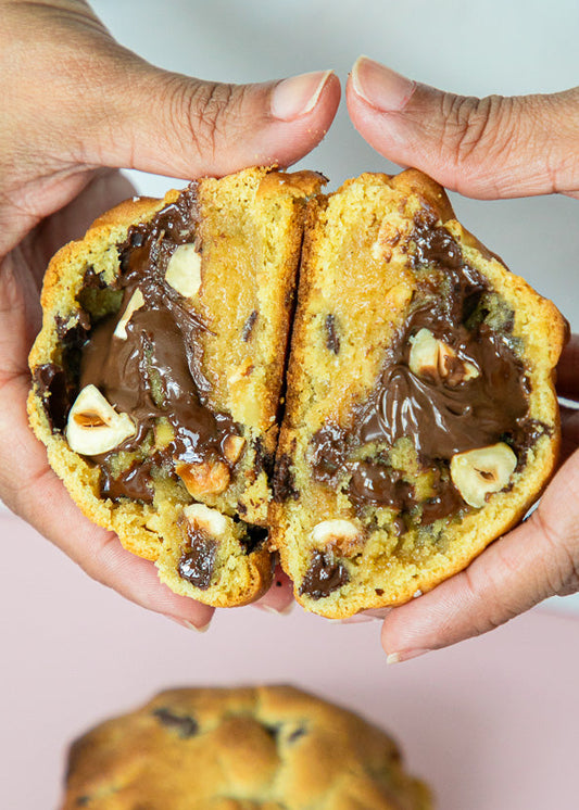 Two hands holding a halved cookie with chocolate filling and hazelnuts, on a light pink background.